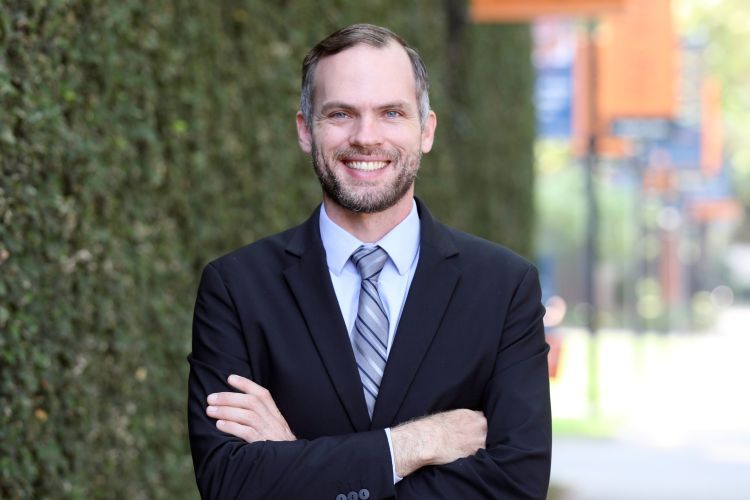 A man in a suit crosses his arm in front of a wall of greenery