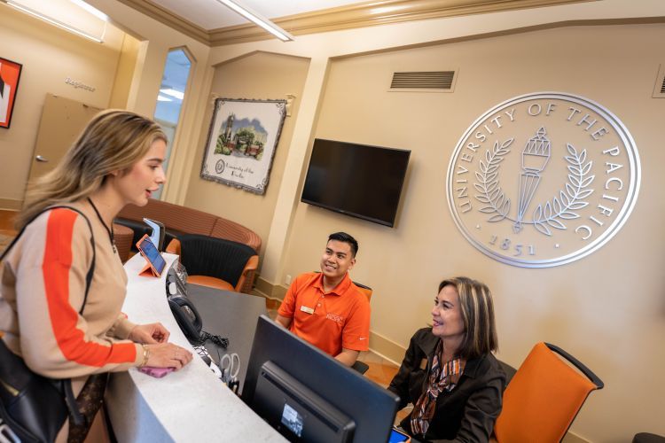 A student stands in front of a desk talking to a student worker and staff member.