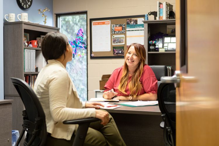 A woman in a salmon shirt seated at a desk smiles at a student