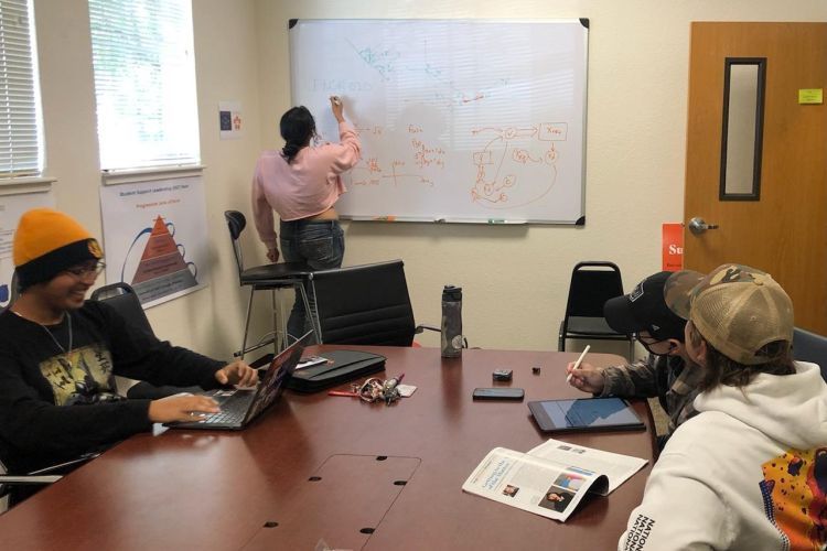 students sit at a table while one student writes on a white board