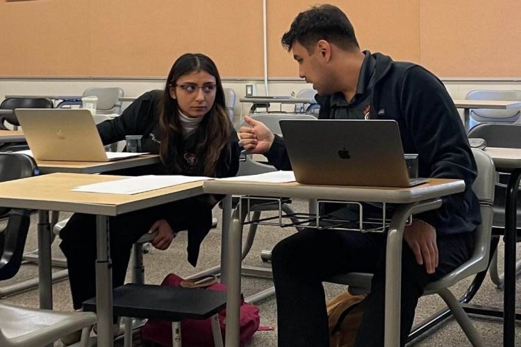 two students sit at desks while talking to each other