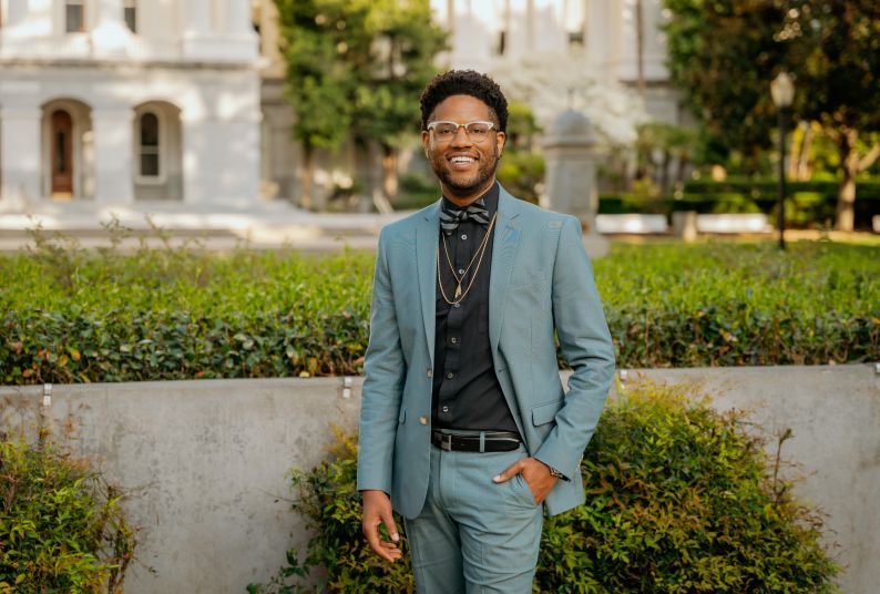 A Black man in a suit standing, smiling, outside the California State Capitol building.