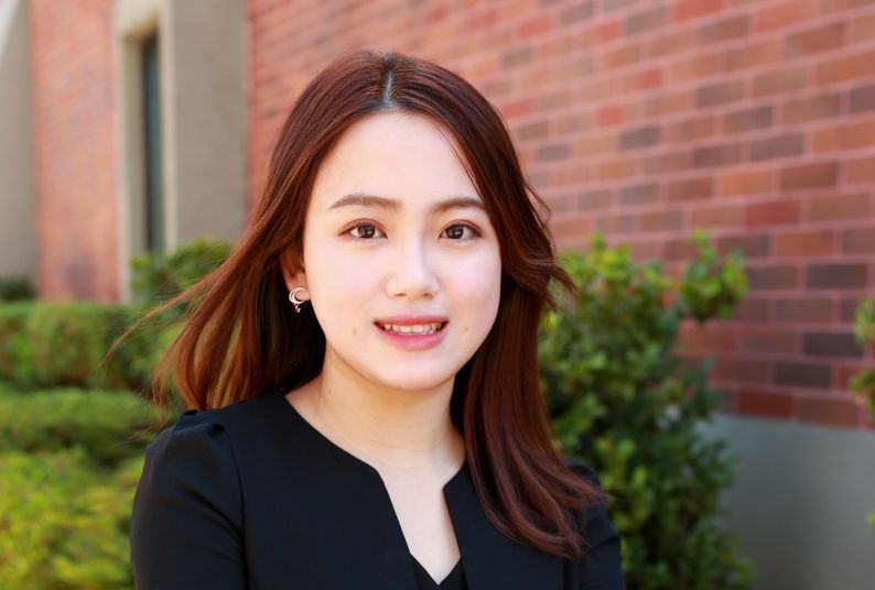 A headshot of an Asian woman in front of a red brick wall