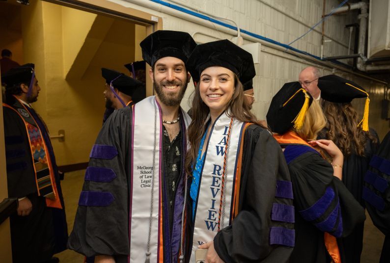 One male individual and one female individual wearing graduation regalia pose for a photo in the basement of an event venue