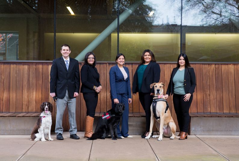 Five people pose in suits in front of a library building, with 3 seated dogs.