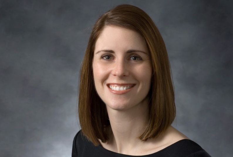 A woman wearing a black top smiles at the camera in front of a neutral gray background 