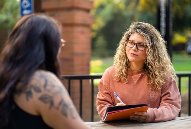 a social work student sits a table holding paperwork while talking to a patient