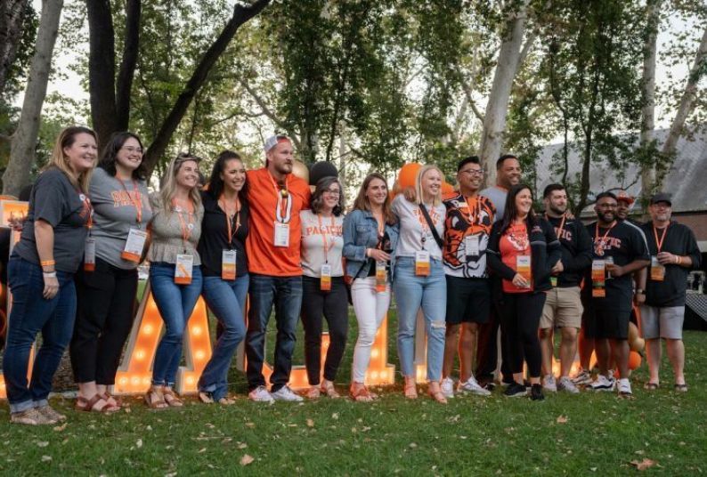 A large group of alumni stand in front of Pacific sign.