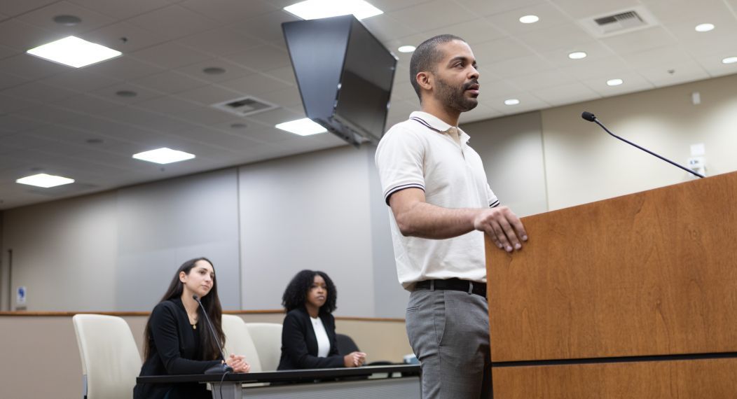 A male student stands at a podium. Two female students are in the background.