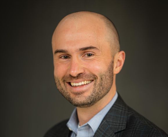 A portrait of a man wearing a suit in front of a gray background