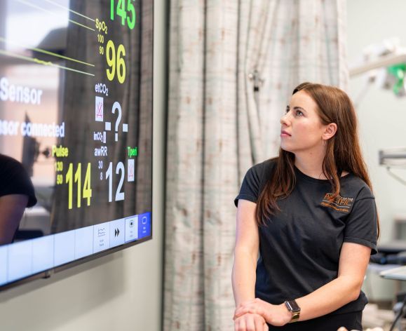 A PA student is shown looking up at a patient monitor. 